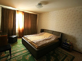 двухцветная прикроватная тумбочка у большой кровати с полосатым покрывалом у стены с белыми цветочными обоями, пуфик и двухцветный гримерный столик у окна светлой спальни