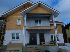 фасад недостроенного элитного двухэтажного дома с полукруглыми ступенями у крыльца и балконом под крышей на зимнем участке