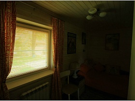закрытые белые жалюзи на окне с цветными красными шторами в спальной комнате с картинами над разложенным диваном деревянной дачи работника кино