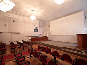 длинный деревянный президиумный стол и деревянная трибуна на полу сцены с большим белым экраном на стене просторного актового зала Дома Культуры времен СССР