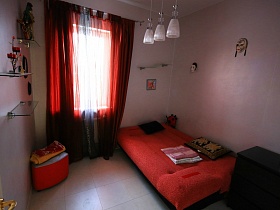 красный разложенный диван с подушками, постельными принадлежностями, красный пуфик у окна с красными шторами в сиреневой спальне художественной дачи в сосновом лесу