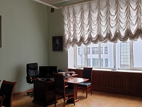 компьютерное кресло у рабочего стола, стулья со спинкой для посетителей у столика рядом в углу светлого строгого кабинета КГБ СССР  с белым ламбрекеном на большом окне для съемок кино