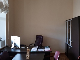 темно коричневый шкаф у стены, высокое кожаное коричневое кресло у рабочего стола секретаря с монитором, блокнотами и бумагами, ксерокс на подоконнике окна приемной при кабинете КГБ СССР для съемок кино