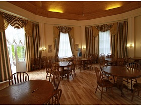 просторный зал летнего кафе с картинами и светильниками на бежевых стенах, двухцветными шторами на высоких окнах, круглыми столиками со стульями на полу с коричневым линолеумом