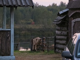 машина на дворе деревянного дома с хозяйственными постройками за открытым деревянным забором на берегу реки большой деревни