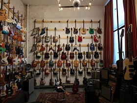 общий вид музыкального магазина с огромным выбором гитар в комнате с белыми стенами, красными шторами на большом окне и цветном ковре на полу