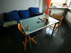 голубой стеклянный стол на круглых ножках у синего мягкого дивана с подушками и сложенным зеленым в клетку покрывалом на современной кухне трехкомнатной квартиры