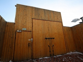 железный засов на высоких воротах для заезда машин и входная дверь в деревянном заборе на территорию банного комплекса