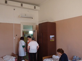 коричневый деревянный шкаф с антресолью за открытой дверью в больничную светлую палату с деревянными кроватями для стационарных больных