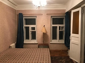 полированная тумбочка, напольный торшер с бежевым абажуром у стены между окнами с белой деревянной рамой и синими шторами на карнизе спальной комнаты с клетчатым матрасом на кровати