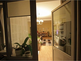 белая люстра с плафонами на белом потолке гостиной над ковром в клетку в зоне отдыха через открытую балконную дверь застекленной лоджии семейной современной квартиры