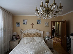 картины и крест на стене над белой деревянной  кроватью с прикраватными тумбочками и большая хрустальная люстра, стилизованная под свечи в светлой спальне большой квартиры врача