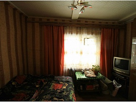 разложенный диван, телевизор на тумбе и столик у окна с красными шторами в спальне дачного домика