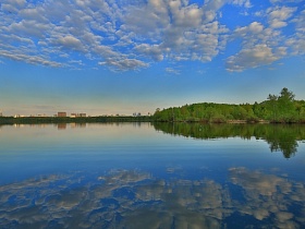 отражение белых облаков на синем небе и сочной зелени деревьев на берегу озера с деревянным причалом