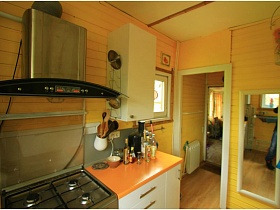 металлическая вытяжка над газовой плитой, встроенной в светлую мебельную стенку, прямоугольное зеркало в белой рамке на деревянной стене у открытой двери на кухню дачного дома