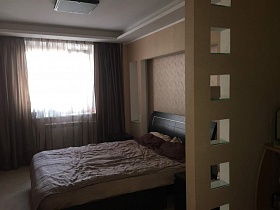 коричневые шторы и белая гардина на окне спальной комнаты с большой кроватью у бежевой стены квартиры с дизайнерским ремонтом