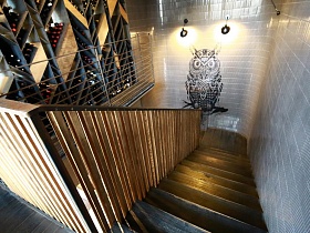 два светильника в стиле хай тек, большая черная сова на серой стене с мозаичной плиткой с одной стороны и винным стеллажом с другой на лестничной клетке ресторана лофт