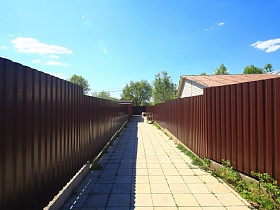 серая квадратная тротуарная плитка на дорожке между высоким коричневым металлическим забором на дачном участке