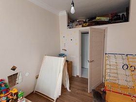 игровой домик, деревянный комод у кремовой стены, разнообразные коробки, ящички и прочие предметы в открытой нише под потолком над открытой дверью детской комнаты квартиры в разных тонах