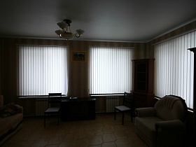 закрытые белые вертикальные жалюзи на окнах бежевой гостиной пустого двухэтажного съемного дома в сосновом лесу