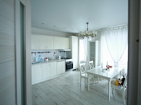 белая кухня, высокий шкаф для посуды со стеклянной дверью, люстра над белым обеденным столом просторной зонированной комнаты из открытой двери спальни квартиры в новостройках