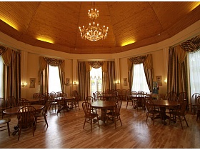 деревянные стулья со спинками вокруг круглых столов на одной ножке по периметру просторного зала уютного летнего кафе с подвесной люстрой по центру высокого купола