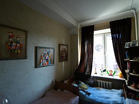 фотографии в рамках, детские игрушки на полках мебельной стенки, на подоконнике окна с коричневыми шторами, яркие картины на стене над кроватками в детской комнате стильной квартиры художника
