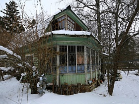 общий вид деревянной советской дачи со старой зеленой краской , многочисленными окнами и шифером на крыше на заснеженном участке с деревьями