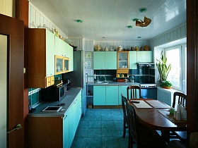 серебристый холодильник между шкафами голубой кухни с бирюзовой квадратной плиткой на полу светлой кухни зонированной комнаты квартиры оператора