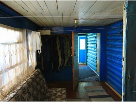 одежда на крючках настенной полки, складной диван у окна с белой гардиной, ковровая дорожка на полу веранды жилого дома в деревне