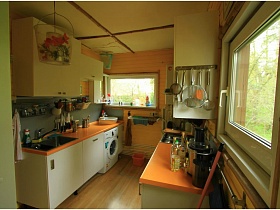 белая мебельная стенка со встроенной мойкой, газовой плитой, стиральной машинкой и оранжевой столешницей на светлой кухне двухэтажной стильной дачи