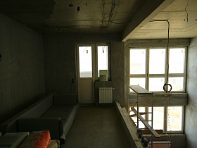 серый раскладной диван у бетонной стены открытой небольшой комнаты на втором этаже с балконной дверью недостроенного пентхауса молодежи для съемок кино