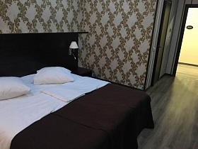 большая двуспальная аккуратно заправленная кровать с коричневым покрывалом, белыми подушками у высокого коричневого изголовья со встроенными розетками в современном номере отеля