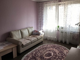 бежевый журнальный столик у светлого мягкого дивана с подушками на полу с овальным цветным ковром просторной гостиной с белой гардиной на окне с балконной дверью современной квартиры молодоженов