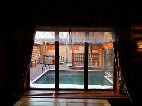 бассейн под открытым небом на территории банного комплекса из окна с цветными шторами домика из сруба