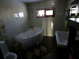 белая раковина на шкафчике, полотенце сушитель,санузел и коврик у белой угловой ванны в комнате с белой плиткой просторного современного дома