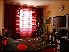 темно красные двухслойные шторы, красные полосатые прозрачные гардины на большом окне восточной спальни с большой кроватью в красных цветах, телевизором и часами на стене