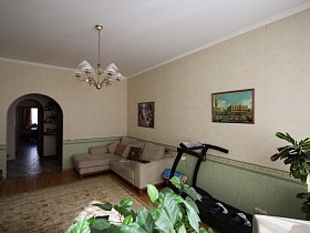 светло серый мягкий угловой диван с подушками, спортивный тренажер,светлый цветной ковер на полу гостиной стандартной четырехкомнатной квартиры на втором этаже жилого дома