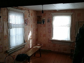 длинная скамейка на деревянном полу комнаты с короткими гардинами на окнах дома заброшенной деревни
