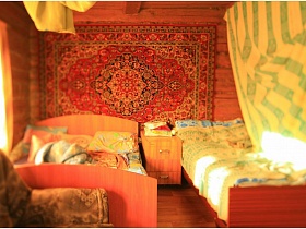 две деревянные кровати,прикроватная тумбочка у стены с цветным ковром в зонированной комнате за желтыми шторами в деревянном жилом доме