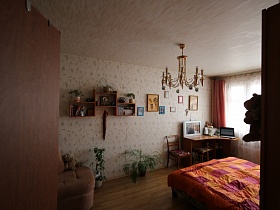 люстра, стилизованная под свечи на светлом потолке, деревянная настенная полка с книгами, картины и фотографии на стене спальной комнаты из открытой двери