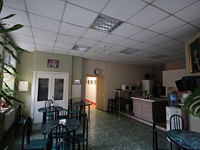 уголок кухни на подиуме, круглые часы над входной дверью в офисное кафе с круглыми уютными столиками на мраморном полу и белым потолком с прямоугольными светильниками