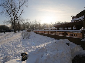 сугробы снега у высокого забора вокруг ресторана в купеческом стиле напротив жилых многоэтажек