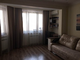 коричневые шторы и белые гардины на больших эркерных окнах гостиной с письменным столом, мягким светлым диваном современной трехкомнатной квартиры