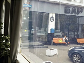 вид на соседний бар, деревянные скамейки у входа, припаркованные машиы через стекла больших окон с серыми шторами кафе