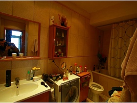 белая раковина со шкафчиком, стиральная машинка, шкаф с туалетными принадлежностями,санузел, шторка над зашитой белой ванной в ванной комнате с желтой плиткой на стенах двухкомнатной квартиры с видом на Москва-сити