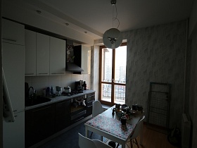 белая с серым мебельная стенка, белый плафон люстры над обеденным столом в современной светлой кухне с прозрачными дверьми в деревянном каркасе на открытый балкон