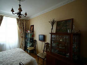 ваза с засушенными цветами, картина,графин и стакан на серванте с посудой,телевизор на стене, два стула с резной деревянной спинкой в спальне с бежевыми стенами советской семейной трехкомнатной квартиры