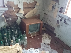 старинный телевизор, пустые стеклянные бутылки на полу неухоженной деревянной комнаты с остатками обоев в доме заброшенной деревни