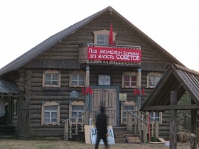 красный плакат, флаг и пятиконечная звезда на перилах балкона двухэтажного деревянного здания конторы с резными наличниками на окнах, деревянными перилами на высоком крыльце